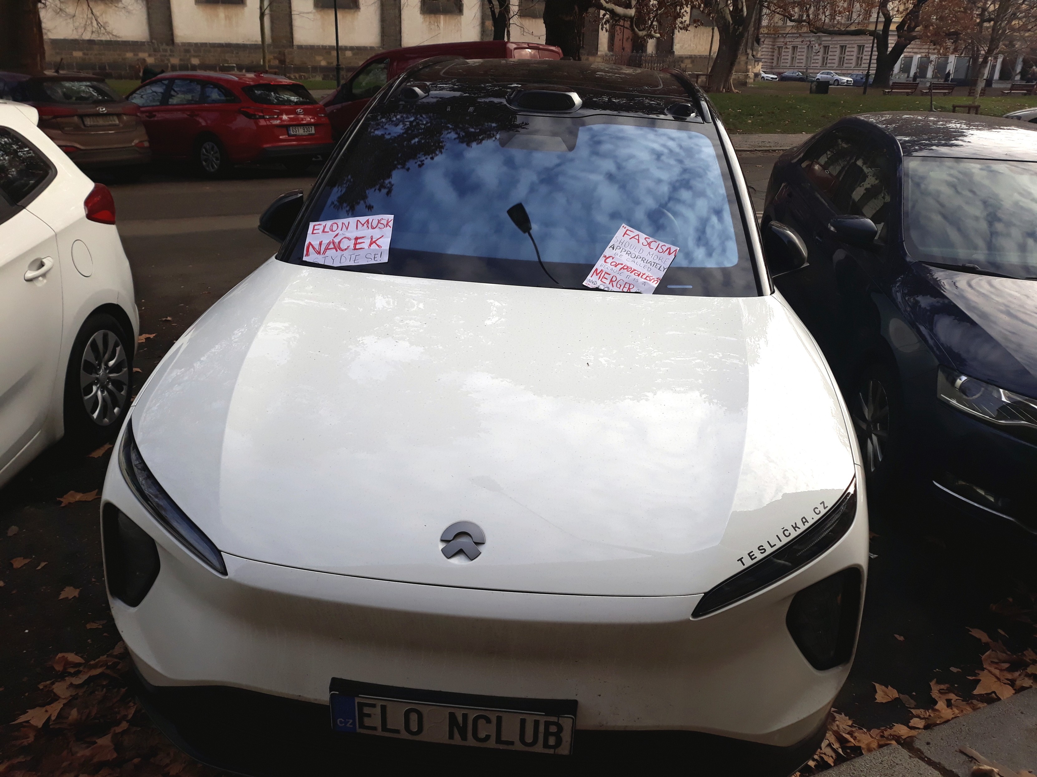 White Nio ET5 EV with licence plate "EL0NCLUB" and teslicka\.cz (luxury Tesla rental business) sticker. Pieces of paper under either windshield say: "Elon Musk je nácek. Styďte se!" and B. Mussolini quote "Fascism \[...\] is a merger of state and corporate power."